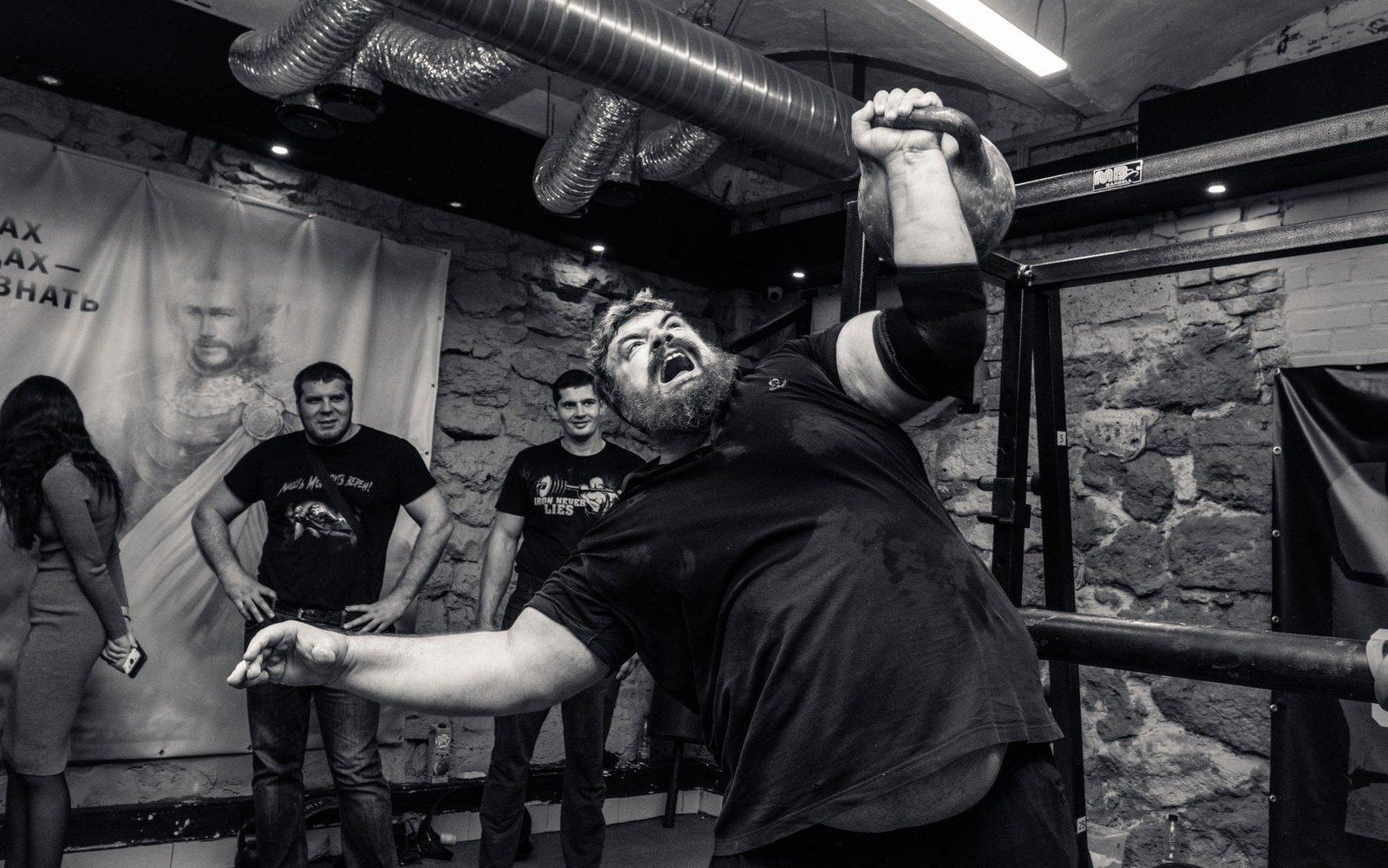 Bearded strongman roars while pressing a kettlebell overhead in a brick-walled gym
