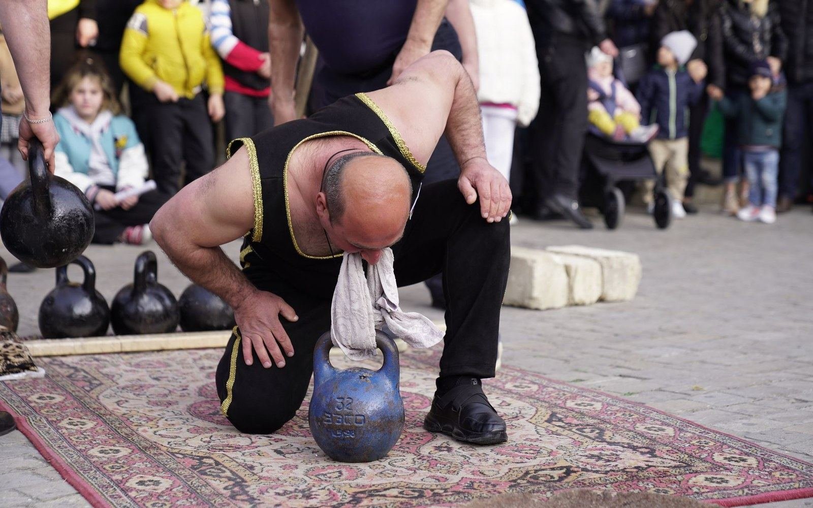A performer lifts a kettlebell using a cloth gripped in his teeth on an outdoor rug