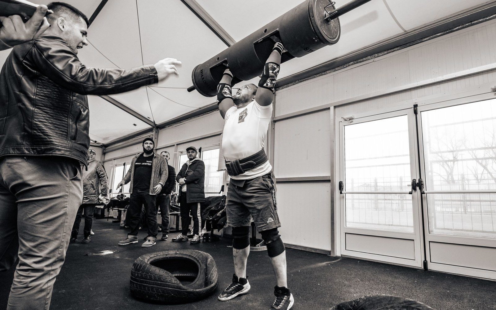 Strongman athlete presses a heavy log overhead as spectators and coaches watch nearby