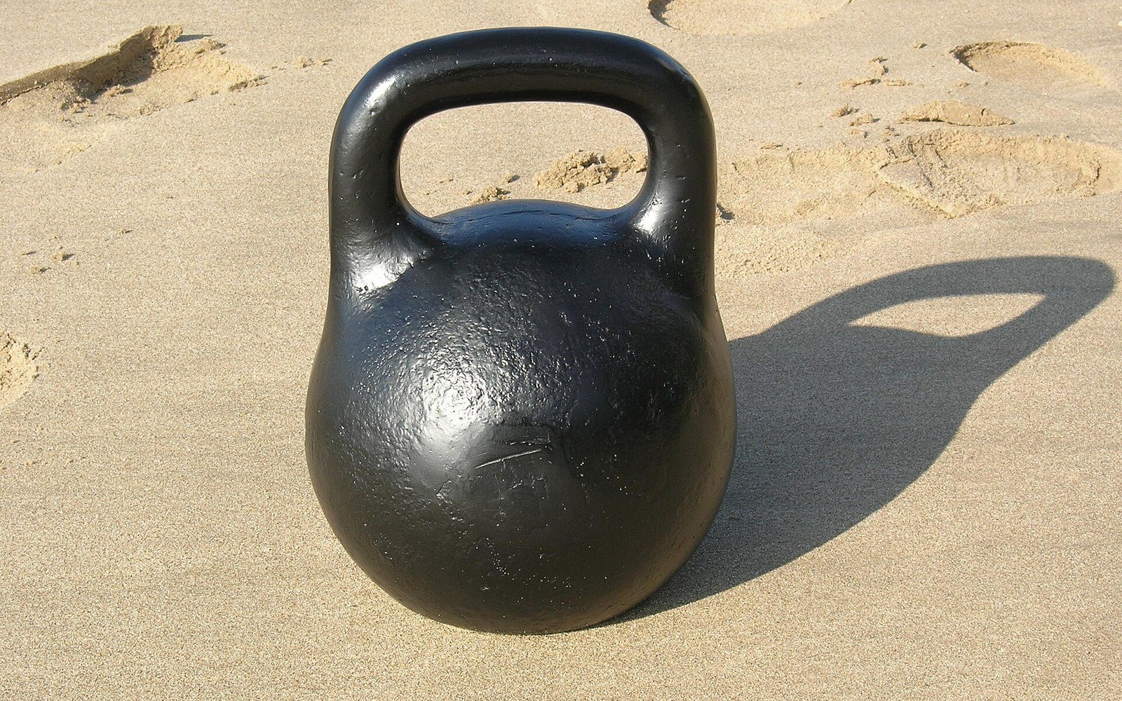 Black cast iron kettlebell resting on sand, casting a long shadow