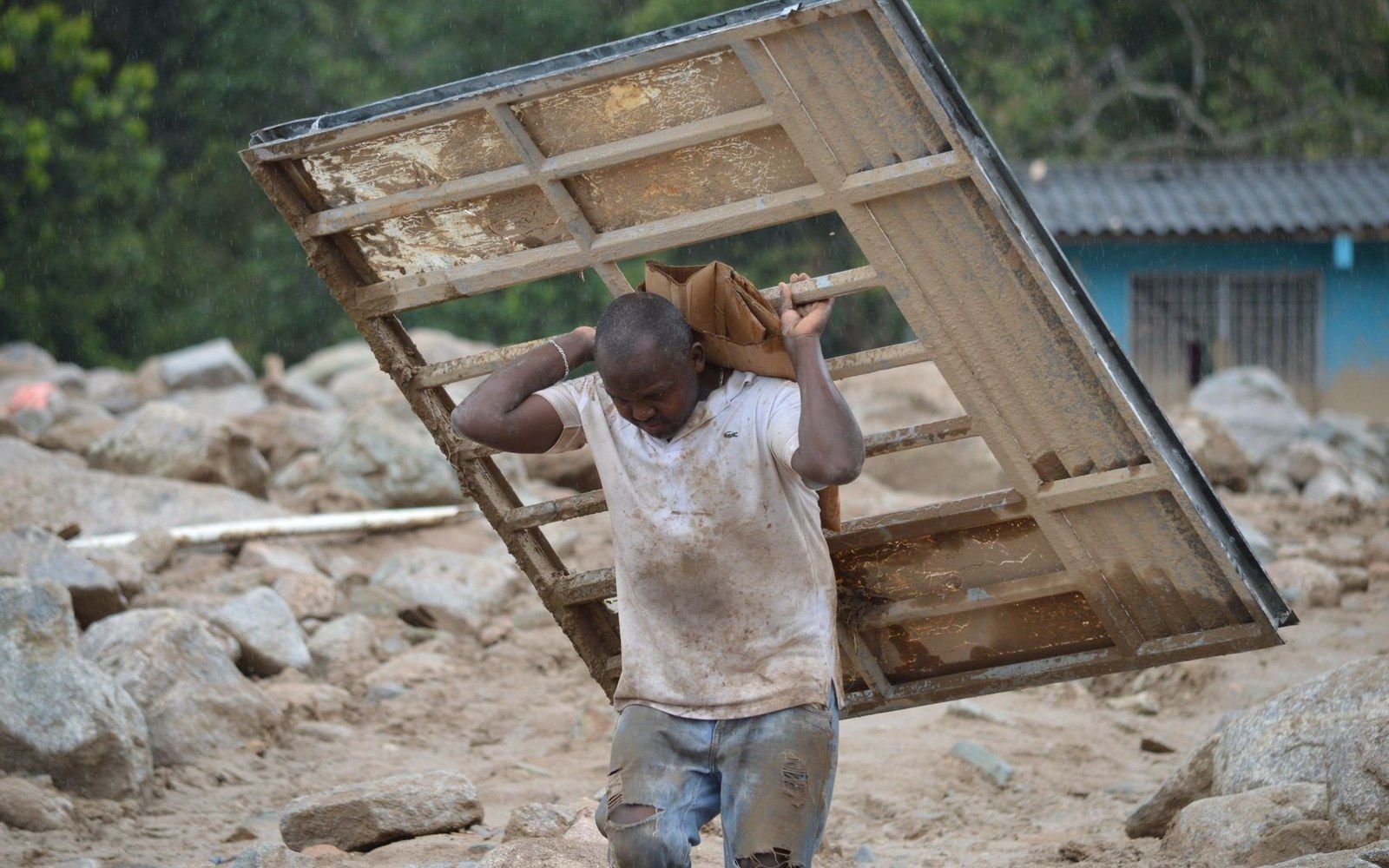 A man carries a mud-caked metal bed frame over debris-strewn ground after flooding