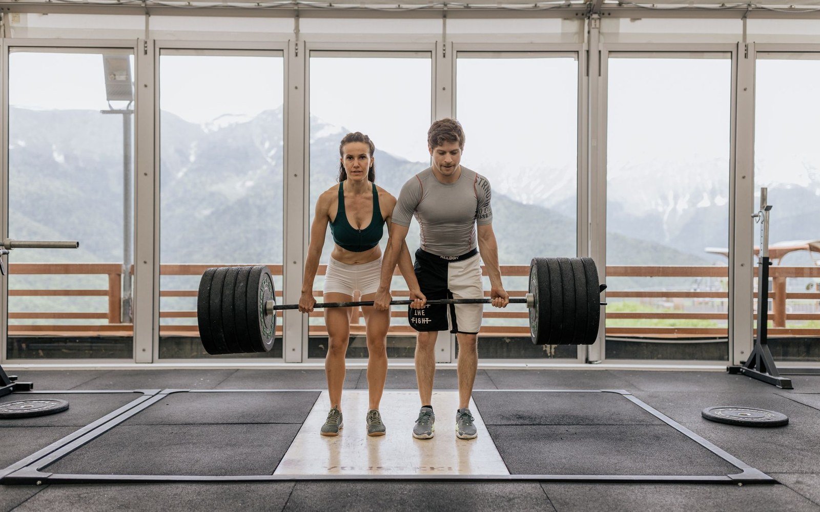 A man and woman deadlift a loaded barbell together in a mountain-view gym