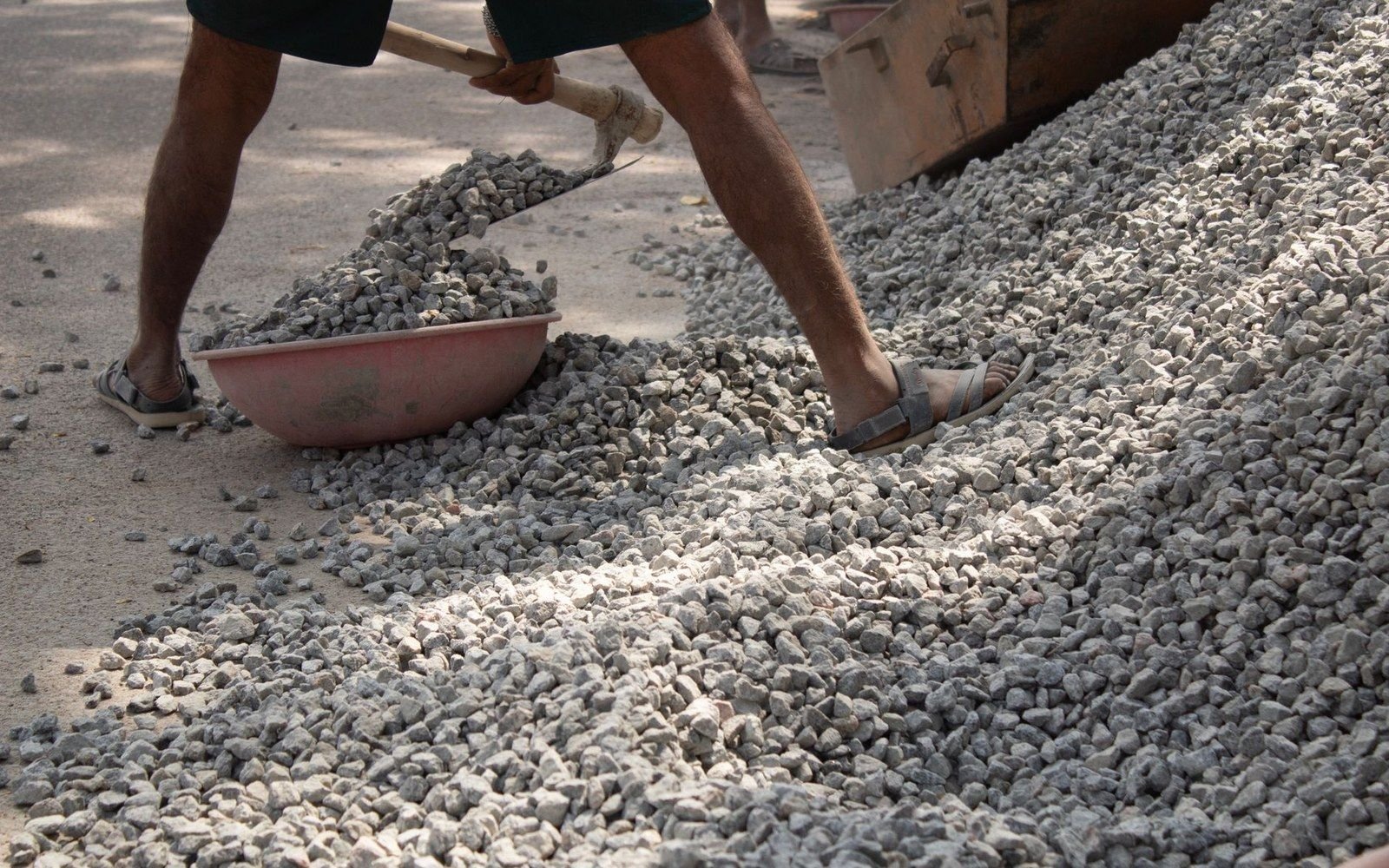 A worker in sandals shovels crushed gravel into a plastic basin on a pile of stones